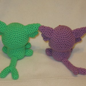 May include: Two crocheted plushies, one green and one purple, with long tails. The green plush has a pointed ear and the purple plush has a rounded ear.