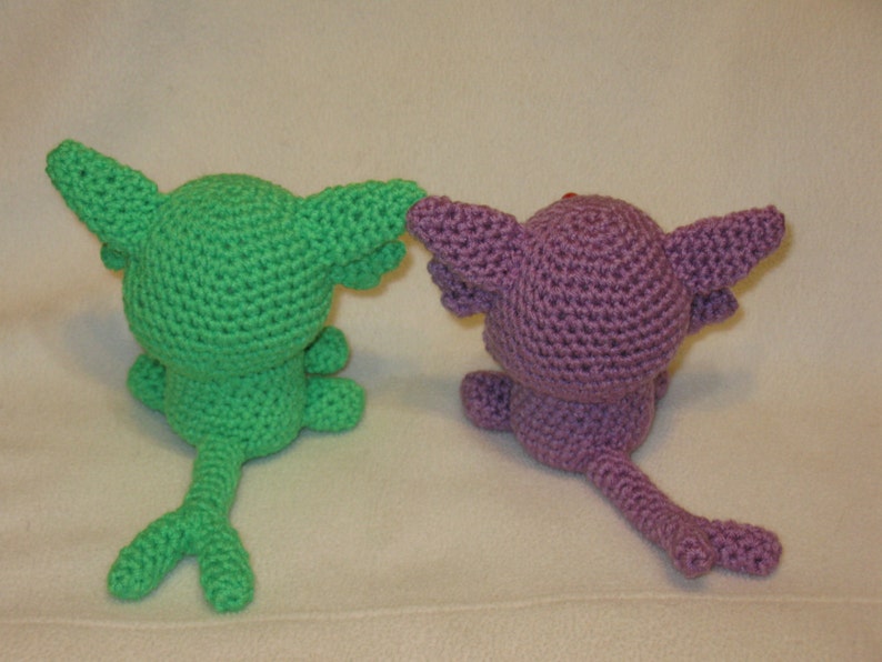 May include: Two crocheted plushies, one green and one purple, with long tails. The green plush has a pointed ear and the purple plush has a rounded ear.