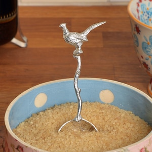 Puede incluir: Un azucarero de cerámica azul y rosa con un diseño floral y lunares blancos. Una cuchara de azúcar de plata con forma de faisán está dentro del tazón.