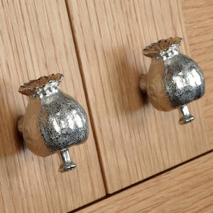 May include: Two silver poppy pod cabinet knobs with a textured finish. The knobs are mounted on a light wood surface.