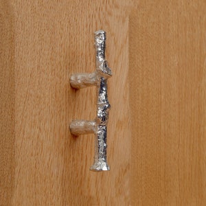 May include: A silver-colored cabinet pull with a bamboo-like design is mounted on a light brown wooden surface. The pull has a vertical stem with horizontal handles on each end, creating a unique and naturalistic aesthetic.