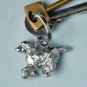 以下が含まれることがあります： 走る犬の彫刻が施された銀色の金属製のキーホルダー。犬は画像の右側を向いており、長い尻尾があります。キーホルダーは銀色のリングと鍵のセットに取り付けられています。