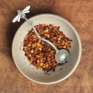 May include: A ceramic bowl filled with red pepper flakes, accompanied by a silver spoon with a dragonfly design. The spoon rests in the bowl, which sits on a wooden surface. The spoon's handle has a twig-like design.