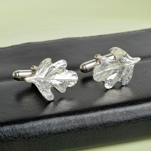 May include: A pair of silver cufflinks featuring an oak leaf design. The leaves are textured and have a detailed, realistic look.