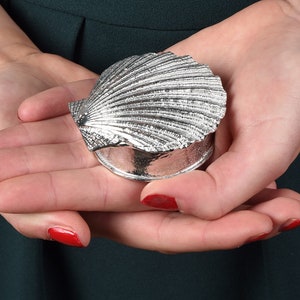 May include: A silver trinket box shaped like a seashell, with a detailed, textured design. The box is closed and resting in the palm of a hand.
