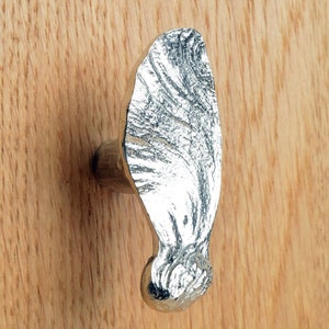 May include: A silver-colored cabinet knob with a textured, organic design. The knob is mounted on a light-colored wooden surface. The knob's shape is elongated with a rounded base and a textured, leaf-like top.