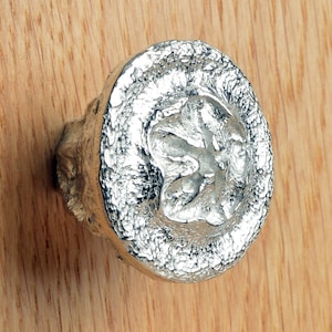 Może przedstawiać: Srebrny metalowy gałka do szafki z fakturowanym, organicznym wzorem. Gałka ma wypukły, stylizowany kształt kwiatu w środku.