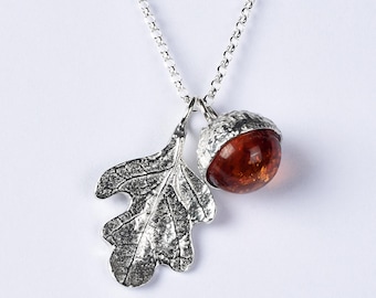 May include: A silver necklace featuring a pendant with a textured silver oak leaf and a round, polished amber acorn. The acorn has a detailed silver cap, and both charms hang from a delicate silver rolo chain against a plain white background.