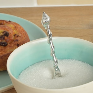 Puede incluir: Una cuchara de azúcar de plata con un tallo retorcido y una parte superior decorativa en forma de flor. La cuchara está apoyada en un cuenco de azúcar blanco.
