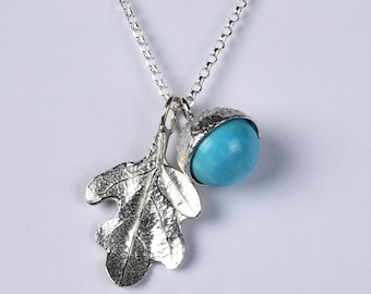 May include: A silver necklace featuring a textured oak leaf charm and an acorn pendant with a bright blue spherical bead. The silver chain is a classic rolo style, connecting to both charms which hang together as a single pendant unit.