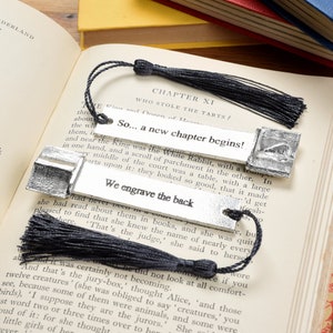 Pode incluir: Dois pingentes de marcador de página de prata com borlas pretas. O marcador de página superior apresenta o texto "So... a new chapter begins!" e o marcador de página inferior apresenta o texto "We engrave the back".
