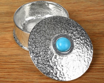May include: A round silver-toned metal trinket box with a textured, hammered finish on the lid and base. The lid features a smooth, circular, bright blue cabochon stone set in the center, contrasting with the metallic, dimpled surface.