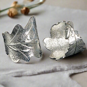 Puede incluir: Dos anillos de servilleta de plata con forma de hojas de roble. Las hojas tienen una superficie texturizada y están detalladas con venas.