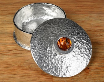 May include: A round silver-toned metal container with a hammered texture. The lid features a circular, polished amber-colored stone set in the center. The interior of the container is smooth and reflective, showing a matching silver-toned finish.