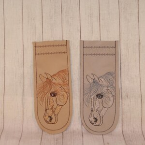 May include: Two beige leather bookmarks with embroidered horse head designs. One horse is brown and the other is gray.