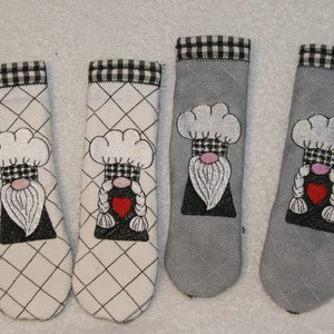 Pode incluir: Quatro pegadores de panela com um design de gnomo. Dois são brancos com acabamento xadrez preto e branco e dois são cinzentos com acabamento xadrez preto e branco. Cada gnomo tem um chapéu de cozinheiro branco, uma barba branca e um coração vermelho.