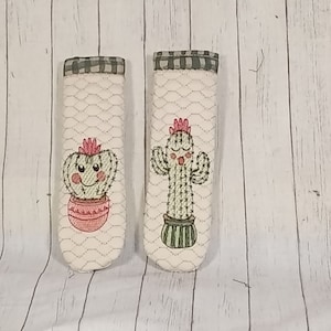 May include: Two white quilted pot holders with embroidered cactus designs. One cactus has a smiling face and the other has a pink flower on top.
