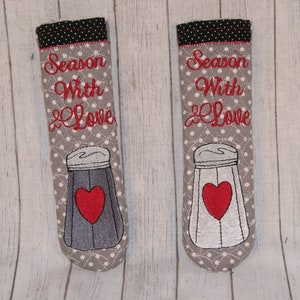 May include: Two gray and white salt and pepper shakers with red hearts. The shakers are embroidered with the text "Season With Love".