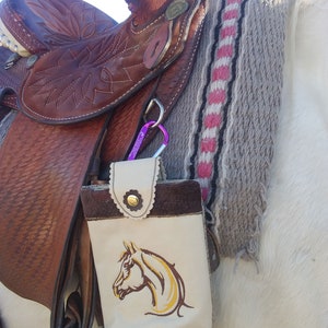 May include: A brown leather saddle with a white and brown embroidered horse head design on a saddle bag. The saddle is on a white horse.