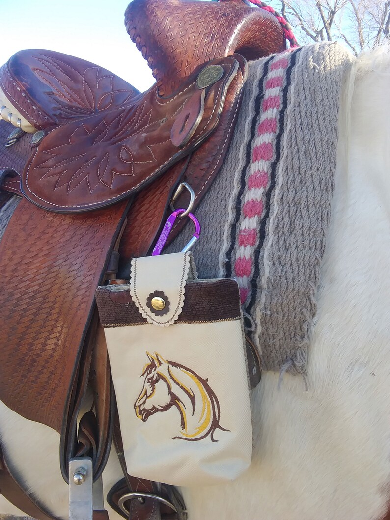 May include: A brown leather saddle with a white and brown embroidered horse head design on a saddle bag. The saddle is on a white horse.