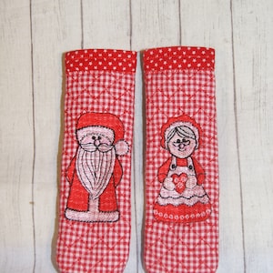 以下が含まれることがあります： サンタクロースとミセス・クロースの刺繍が施された、赤と白のキルト製の鍋つかみ2枚セット。鍋つかみはギンガムチェック柄で、赤い星で縁取られています。