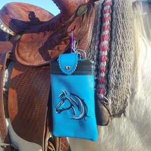 May include: A blue leather phone case with a horse head design attached to a saddle on a white horse.
