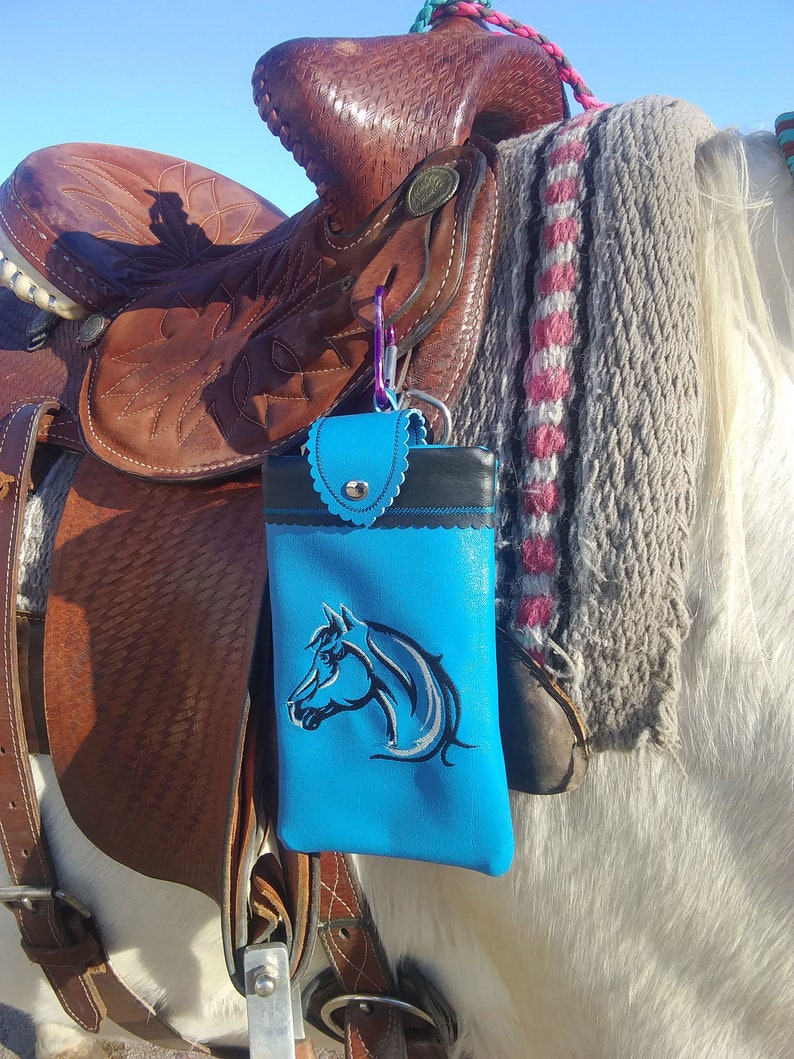 May include: A blue leather phone case with a horse head design attached to a saddle on a white horse.