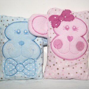 May include: Two plush toy animals, one blue with a bow tie and one pink with a bow, both with brown polka dots on a white background.