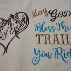 May include: Embroidered white fabric with two brown horses in a heart shape and the text "May God Bless The Trail You Ride" in brown and blue cursive.