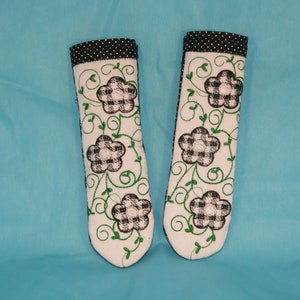 May include: A pair of white pot holders with black and white checkered flowers and green vines embroidered on them. The pot holders have a black and white polka dot trim.