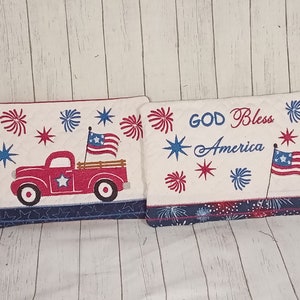 Puede incluir: Dos toallas de cocina bordadas en rojo, blanco y azul. Una toalla presenta un camión rojo con una bandera en la parte trasera y la otra toalla presenta el texto "God Bless America" con una bandera en el lado derecho. Ambas toallas tienen un borde azul y blanco con estrellas y fuegos artificiales.