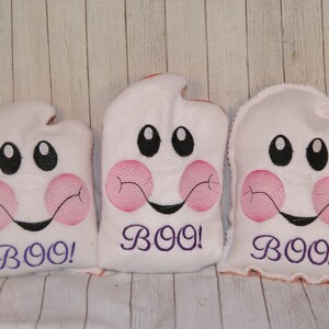 May include: Three white plush ghost pillows with pink cheeks and embroidered black eyes. Each pillow has the word "BOO!" embroidered in purple on the front.