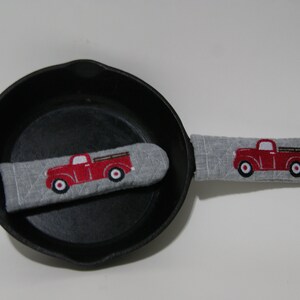 May include: A grey fabric pot holder with a red truck design. The pot holder is on a black cast iron skillet.