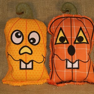 May include: Two handmade orange and yellow fabric pumpkins with stitched-on faces. One pumpkin has a plaid pattern and the other has a repeating pattern of small orange flowers. Both pumpkins have black eyes, a black nose, and white teeth.