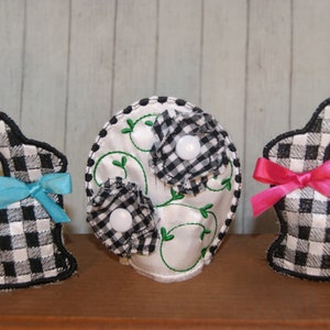 May include: Three black and white checkered fabric Easter bunny ornaments with bows. The bunnies are standing on a wooden surface. The middle bunny is holding a white fabric flower with green leaves.