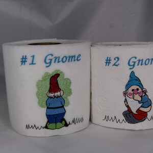May include: Two white toilet paper rolls with embroidered gnomes. The first roll has the text "#1 Gnome" and the second roll has the text "#2 Gnome".
