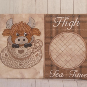 May include: Embroidered tea cozy featuring a Highland cow in a teacup. The design includes the words "High Tea Time" in elegant script. The cozy is made of tan and brown plaid fabric, with a heart detail on the teacup.