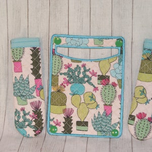 May include: A set of three fabric items featuring a cactus design. The set includes a wallet and two matching pen holders. The items have a light pink background with various cacti in pots, outlined in green and blue.