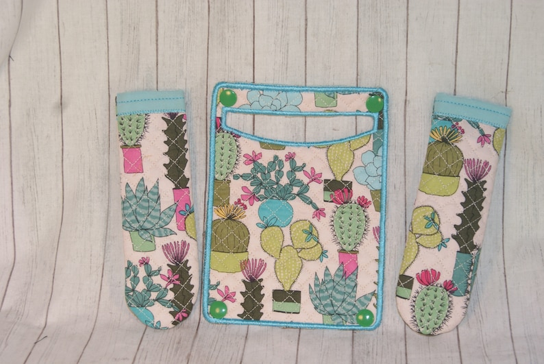 May include: A set of three fabric items featuring a cactus design. The set includes a wallet and two matching pen holders. The items have a light pink background with various cacti in pots, outlined in green and blue.