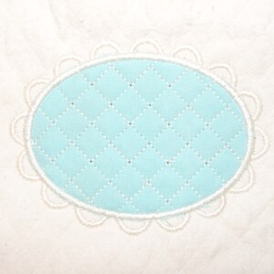 May include: A close-up of a white fabric with a light blue oval-shaped patch. The patch has a quilted pattern and is surrounded by a white scalloped border. The fabric has a subtle texture.