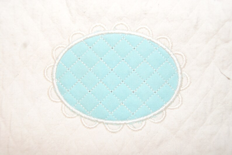 May include: A close-up of a white fabric with a light blue oval-shaped patch. The patch has a quilted pattern and is surrounded by a white scalloped border. The fabric has a subtle texture.