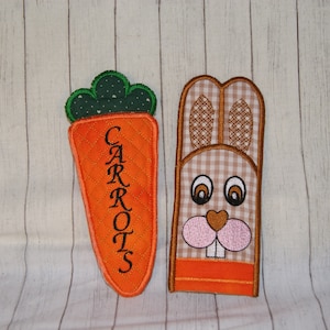 Può includere: Decorazioni per la cucina ricamate a forma di carota e coniglietto. La carota è arancione con foglie verdi e la parola "CARROTS" in nero. Il coniglietto ha un motivo a scacchi e un naso rosa.