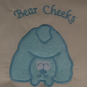 Può includere: Un disegno ricamato di un orso blu chiaro con il testo "Bear Cheeks" sopra.