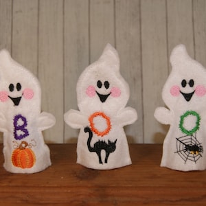 Puede incluir: Tres títeres de dedo de fieltro blanco con forma de fantasmas con caras bordadas. El primer fantasma tiene una "B" morada y una calabaza naranja. El segundo fantasma tiene un círculo naranja y un gato negro. El tercer fantasma tiene una corona verde y una araña en una telaraña.