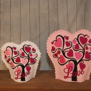 Puede incluir: Dos adornos de fieltro en forma de corazón con fondo rosa y blanco. Cada adorno presenta un árbol negro con corazones rosas y rojos. La palabra "Love" está bordada en la parte inferior de cada adorno.