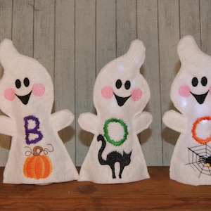 Puede incluir: Tres adornos de fantasmas de fieltro blanco con caras y detalles bordados. El primer fantasma tiene una "B" morada y una calabaza, el segundo tiene una "O" verde y un gato negro, y el tercero tiene una "O" naranja y una telaraña.
