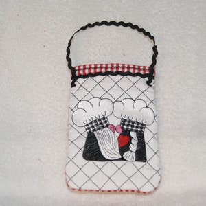 May include: A white fabric bag with a black handle and red and white checkered trim. The bag has an embroidered design of two chefs wearing hats and holding a heart.