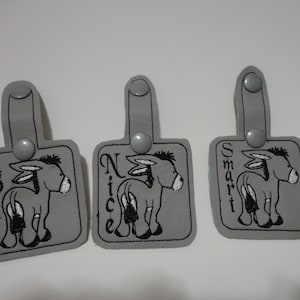 May include: Three gray fabric keychains with a donkey design and the words "Bad", "Nice", and "Smart" embroidered on them.