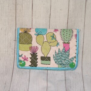 May include: A fabric wallet featuring a cactus print in shades of green, pink, and blue on a cream background. The wallet is quilted with a turquoise border and green button closures. The design includes various cactus illustrations.