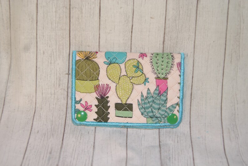 May include: A fabric wallet featuring a cactus print in shades of green, pink, and blue on a cream background. The wallet is quilted with a turquoise border and green button closures. The design includes various cactus illustrations.